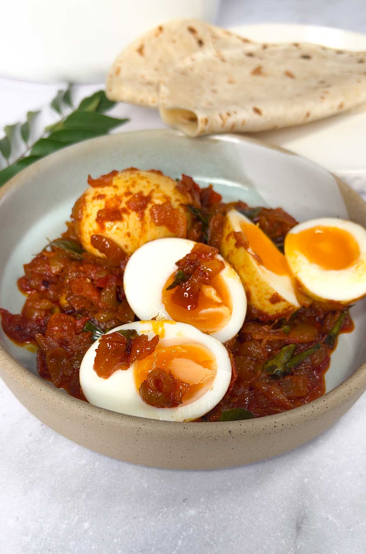 Kerala Egg Roast in a plate with chapathi in the background. A dish of boiled eggs in a lightly spiced, caramelised onion & tomato thick sauce.