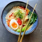miso noodle soup in a blue bowl, with golden chopsticks on top. Topped with a soft boiled egg, blanched enoki mushrooms and boy choy, chilli oil, spring onion, red chillies
