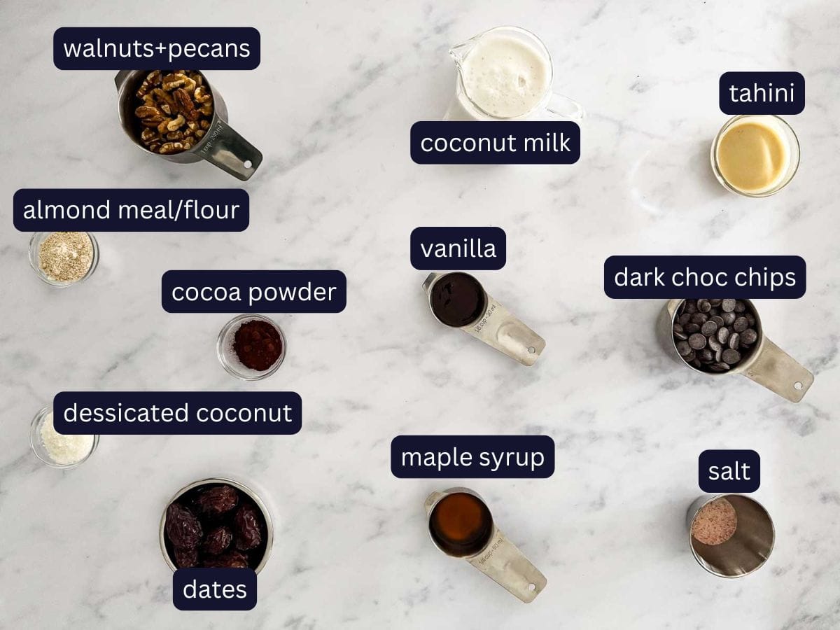Flat lay of labelled ingredients for healthy chocolate mousse bars, including walnuts, pecans, dates, coconut milk, tahini, dark chocolate chips, cocoa powder, vanilla, almond flour, desiccated coconut, maple syrup, and salt.