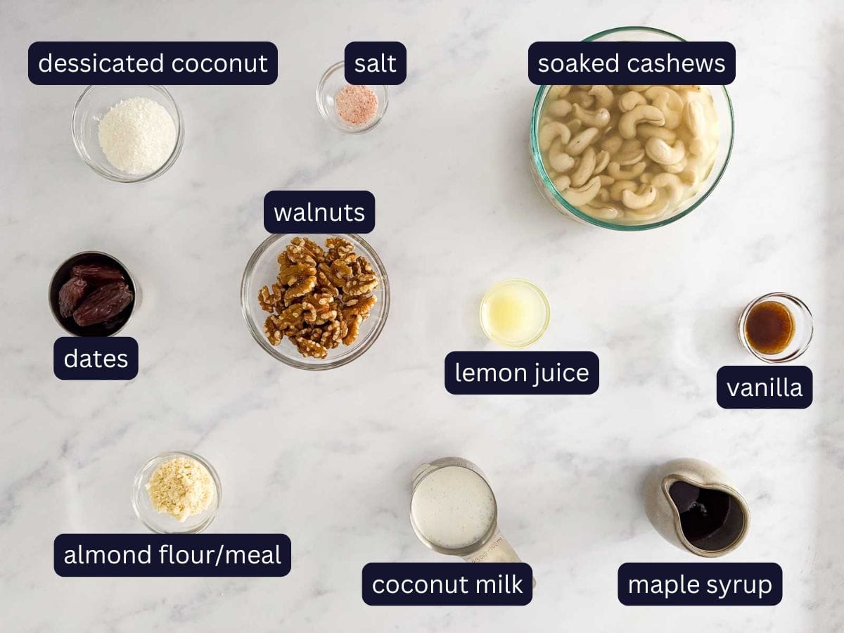 ingredients to make vegan no bake cheesecake from top view on marble background. ingredients include, desiccated coconut, salt, dates, soaked cashews, walnuts, lemon juice, vanilla, almond flour, coconut milk and maple syrup