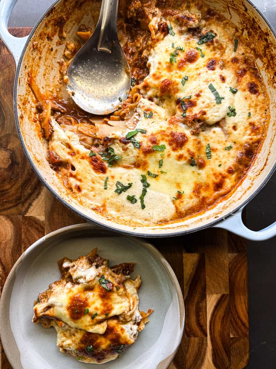 cooked lasagna in baby blue dutch oven skillet with spoon inside. a small plate beside it with a piece of lasagna on plate. wood background.