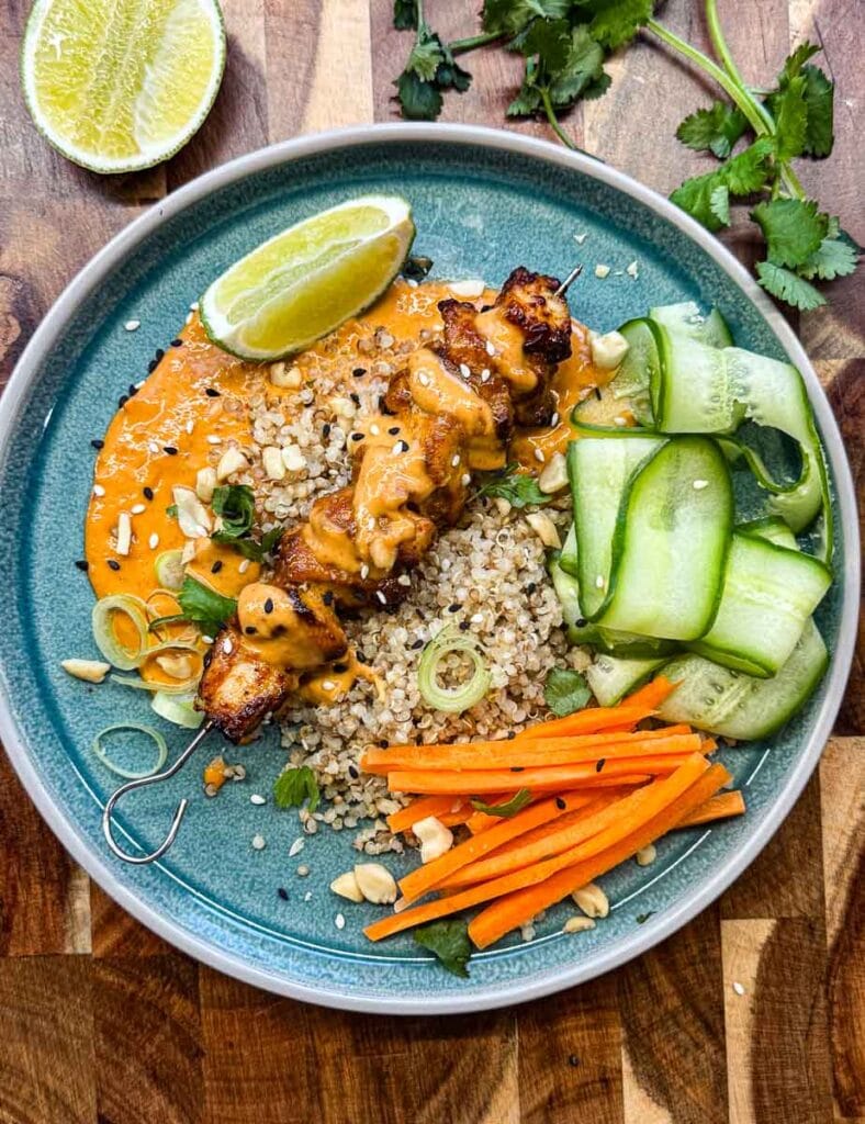 satay chicken skewer on bed of quinoa, on a blue plate with wedge of lime, cucumber ribbons, carrot