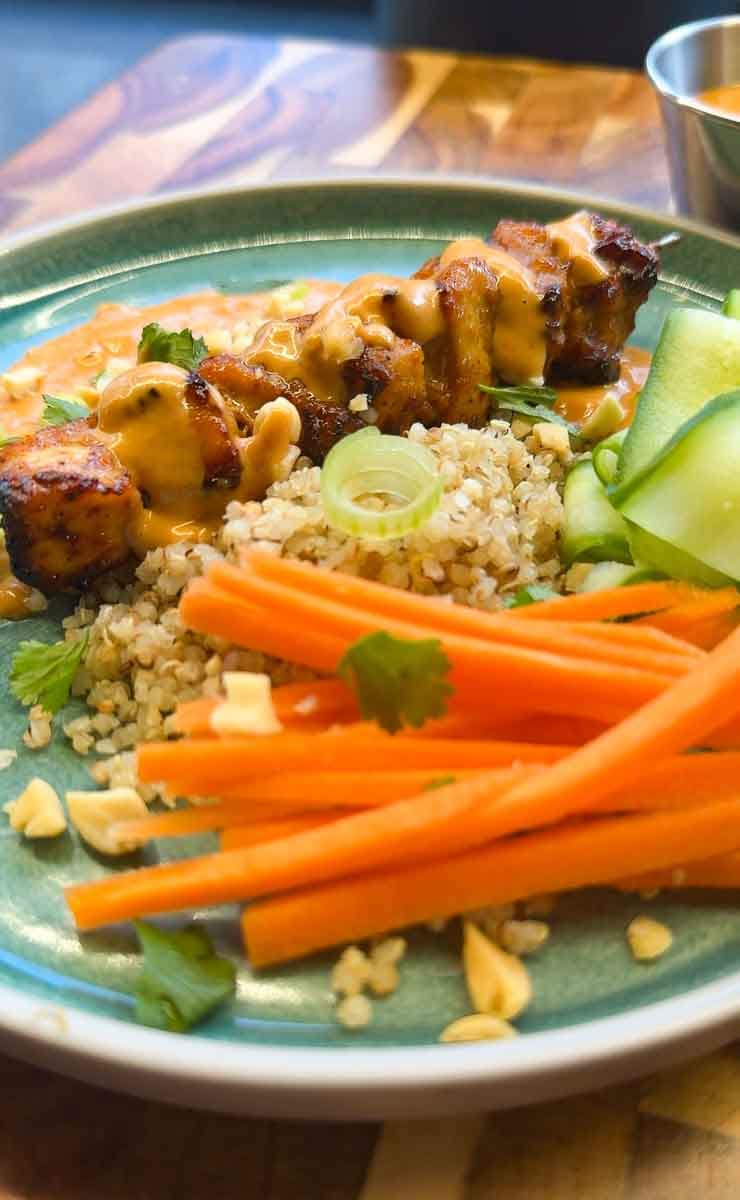 high-protein air fryer satay chicken skewers served with creamy peanut sauce, fluffy quinoa, fresh carrot, cucumber ribbons, spring onion and chopped peanuts on a green plate.