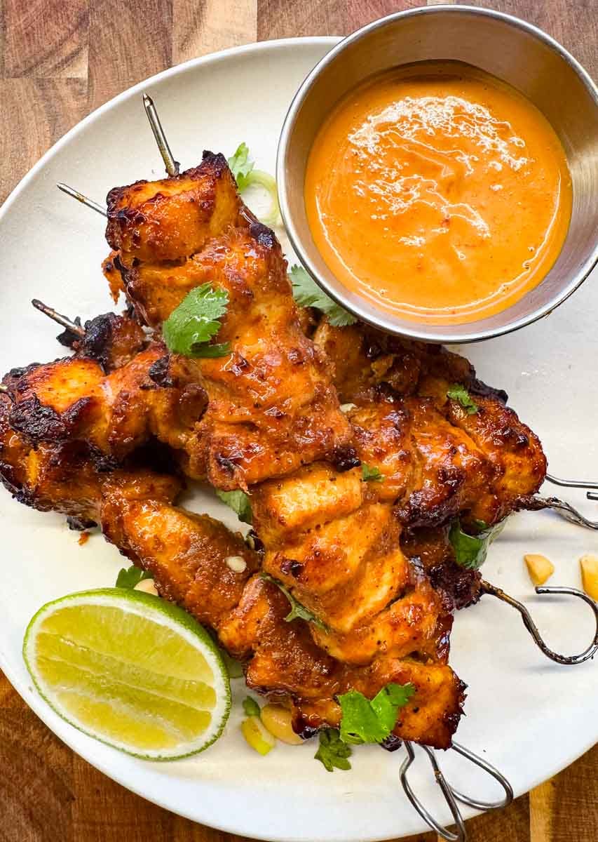 air fryer satay chicken skewers served on a white plate, with creamy homemade peanut sauce in a small steel bowl and a fresh wedge of lime. garnished with coriander.