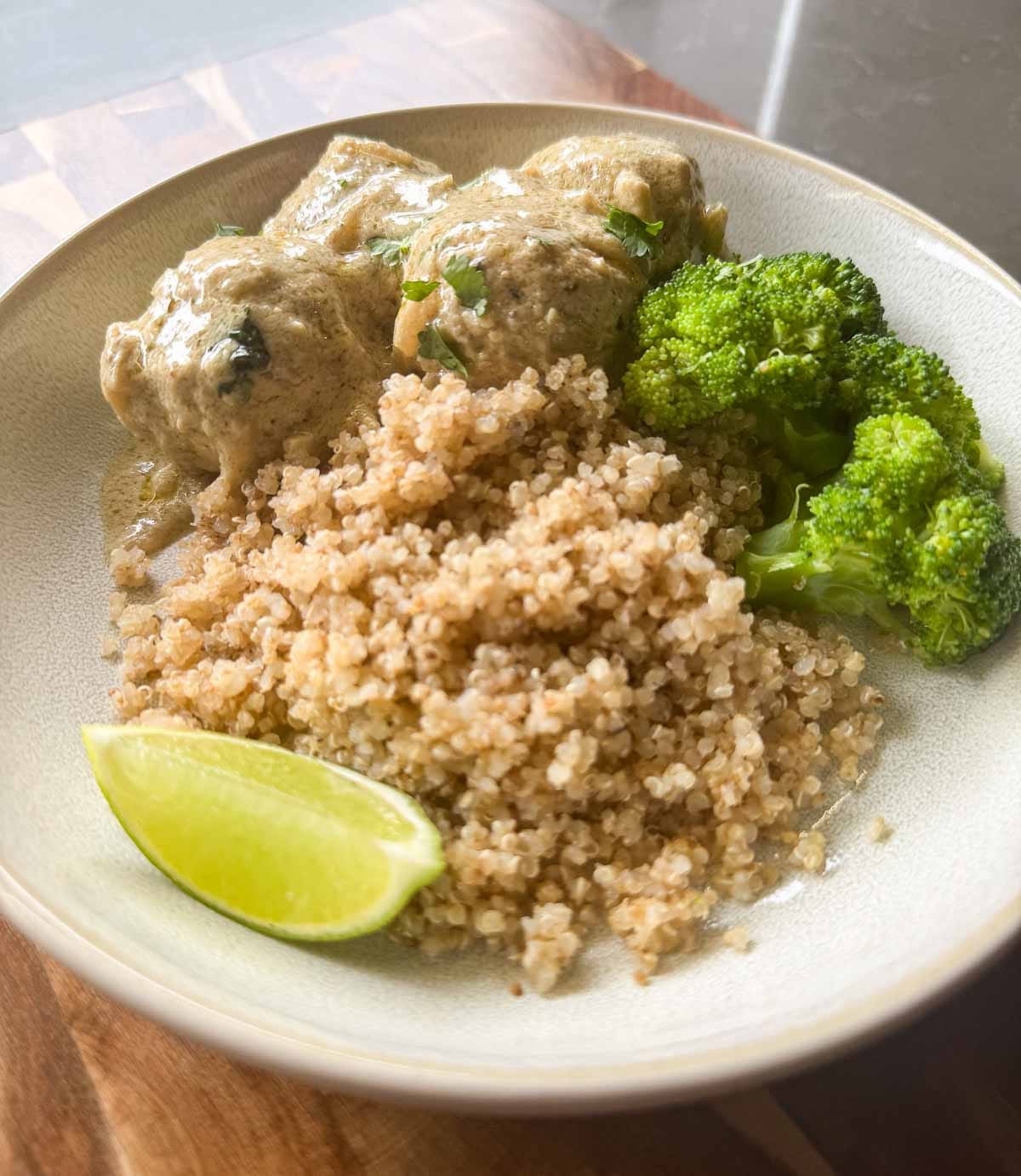 a off white plate, with creamy chicken kofta curry, quinoa, a wedge of lime and some steamed broccoli.