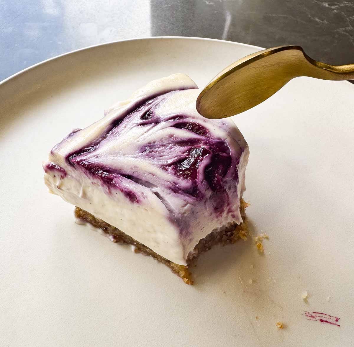 slice of Vegan no bake cheesecake on white plate,  that has been eaten into with a golden spoon.